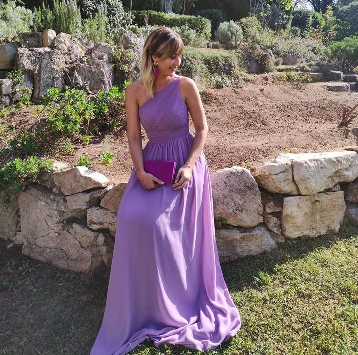 una invitada de boda sentada en un muro con un vestido largo de gasa en color lila de escote asimétrico